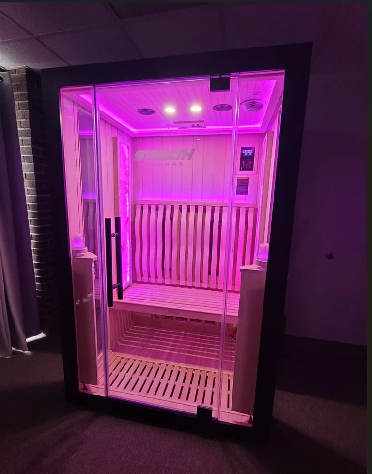 A modern infrared sauna cabin with neon pink lighting, glass door, wooden interior benches, and LED lights mounted on the ceiling