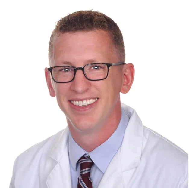 Professional headshot of a smiling man wearing glasses, white lab coat, and striped tie against white background